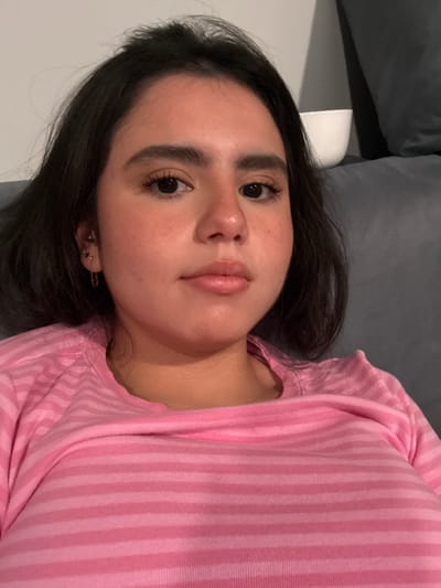 A young woman is shown from the chest up, with her face turned slightly towards the camera. She has dark hair, dark eyes, and appears to be wearing light makeup. She is wearing a pink striped shirt. The background is blurry and appears to be an indoor setting, possibly a couch or sofa. No specific Austin landmarks or context are visible.