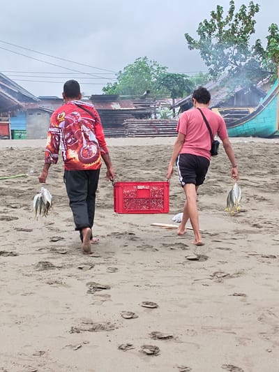 Two individuals are walking on a sandy beach in Curehtunong, Indonesia. The person on the left is wearing a long-sleeved red shirt with a motorcycle graphic and black pants, carrying a bunch of fish in their left hand and a red crate in their right. The person on the right is wearing a pink t-shirt, black shorts with white stripes, and carrying a bunch of fish in their left hand. In the background, there are several small buildings with corrugated metal roofs, some wooden planks, a green boat, and lush green trees. The sky is overcast.