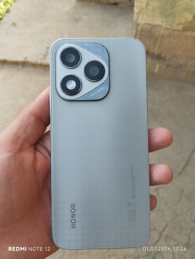 A person's hand holds a silver smartphone, revealing its rear design. The phone features a prominent camera module with three circular lenses arranged in a triangular formation. Text on the camera module reads "108MP ASPH". The "HONOR" brand name is visible vertically on the lower left side of the phone's back. Near the bottom right, regulatory marks like "CE" and "UKCA" are present, along with "Honor Device Co. Ltd Shenzhen, 518040 P.R.C" and "Made in China Model ABR-NX1". A timestamp "01.07.2026 17:26" is overlaid on the image. The background consists of textured, light-colored paving stones and a blurry, sandy-colored wall or structure, suggesting an outdoor setting. The lighting appears natural, indicating daytime. The focus is on the smartphone, with the hand and background being less detailed.