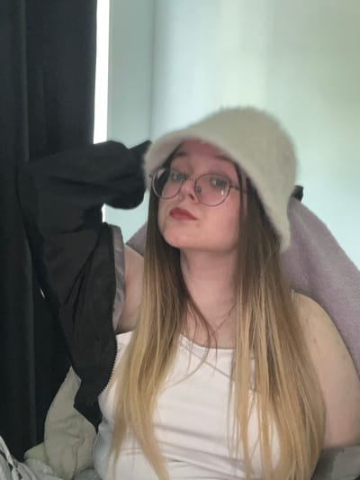 A young person with long, light brown hair is wearing a white, furry hat and round-framed glasses. They have fair skin and red lipstick. They are wearing a white shirt under a black jacket. The person is sitting on a black surface, possibly a couch. A grey or lavender blanket is partially visible in the background. The location is not identifiable as Pushchino, Russia. The image is oriented sideways.