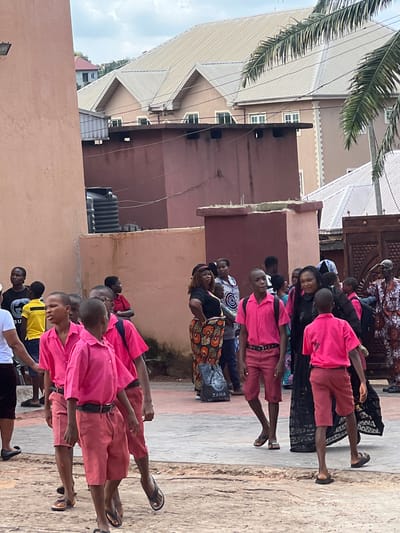 The image depicts an outdoor scene in the city of Nnewi, Nigeria, likely on a street or within a school compound. In the foreground, several young boys are walking, all wearing matching pink short-sleeved shirts and coral-colored shorts. Most of the boys are wearing black belts. Some have backpacks. To the left, an adult woman is partially visible, wearing a white t-shirt and dark shorts. Behind the boys, a group of adults and children are gathered. The architecture in the background suggests a residential or institutional setting, with buildings featuring corrugated metal roofs and decorative facades. There are palm trees and wires visible, indicating a tropical or sub-tropical environment. The ground appears to be paved with concrete and dirt. The overall atmosphere seems to be one of daily activity, possibly during a school day or an event.