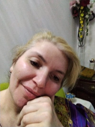 A woman with blonde hair, resting her chin on her hand, is seen in a close-up selfie. She is looking towards the camera with a slight smile. The background is softly lit and shows a wall with a floral arrangement in a vase and a wooden dresser. There is no indication of the city of Al Mas'oudi, United Arab Emirates.