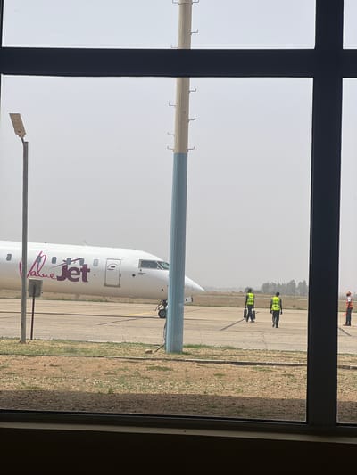 From an elevated vantage point, likely within a building, the scene depicts an airport tarmac viewed through a multi-pane window. In the foreground, a ValueJet-branded aircraft, likely a Bombardier CRJ900 based on size and shape, is parked. Airport ground crew members wearing high-visibility vests are visible near the plane and further away along the tarmac. The environment appears hazy or dusty. This location can be identified as Nnamdi Azikiwe International Airport in Kwi, Nigeria, due to the presence of ValueJet aircraft, a Nigerian domestic airline.