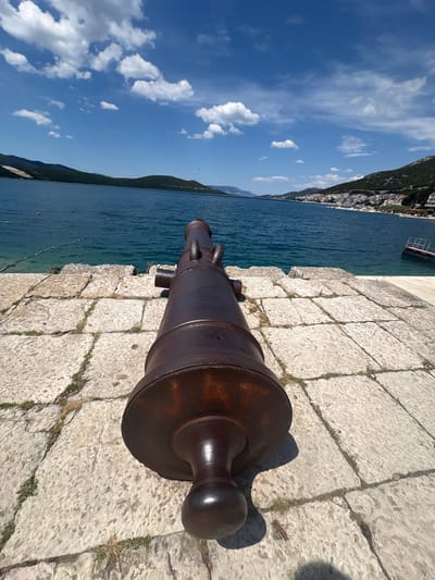 A cannon is positioned on a stone platform overlooking the Adriatic Sea in Neum, Bosnia and Herzegovina. The cannon is made of metal, possibly iron or bronze, and appears to be historic. It is positioned at the edge of the platform, facing out towards the water. The stone platform is constructed from large, rectangular blocks. The sea is calm and blue. A mountain range and the city of Neum are visible along the coastline. The sky is blue with scattered clouds.