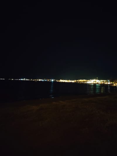 The image is a night-time, long-shot photograph taken from a beach, looking out over a body of water towards a brightly lit city skyline. The sky is completely dark. The water in the foreground is dark and reflects some of the city lights, creating streaks of illumination on its surface. The beach in the immediate foreground is dark and indistinct, appearing as a shadowed area of sand or ground. The city lights stretch horizontally across the horizon in the distance, forming a continuous line of white, yellow, and orange glows. These lights suggest buildings and structures illuminated for the night. The water appears calm, with a subtle ripple. There are no visible people or distinct objects in the scene, only the broad strokes of the illuminated cityscape meeting the dark expanse of the sea and sky. The location is identified as a coastal area in Unknown, Egypt.