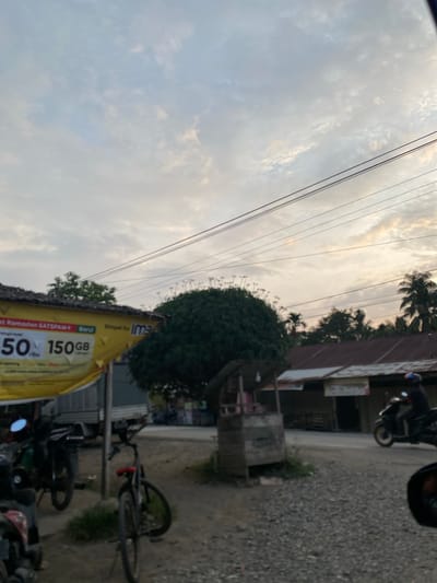 The image depicts a street scene in Uleejalan, Indonesia, during what appears to be either dawn or dusk, with a cloudy sky. In the foreground, a gravelly road is visible. To the left, a yellow banner advertising a mobile data package with "50" and "150GB" is partially obscuring a parked motorcycle and a truck. A bicycle is parked in the middle ground. Behind these objects, a small wooden kiosk stands on the side of the road, with a large, dense tree behind it. Further back, buildings with corrugated metal roofs line the street, and a person is riding a motorcycle towards the right side of the frame. Palm trees and other foliage are scattered throughout the background, indicating a tropical or subtropical environment. Power lines are strung across the upper portion of the image.
