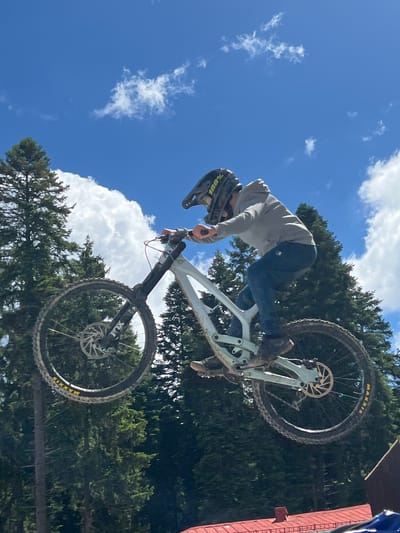 A mountain biker is captured mid-air, performing a jump against a backdrop of a bright blue sky with scattered white clouds and a dense forest of tall evergreen trees. The biker, wearing a helmet and a grey hoodie with blue jeans, is positioned on a light-colored downhill mountain bike, its wheels and suspension clearly visible. The bike has "MAXXIS" branding on the tires and "FOX" on the front fork. The surrounding environment suggests an outdoor recreational area, possibly a bike park or a natural trail, indicated by the presence of trees and a partial view of a red-roofed structure at the bottom right of the frame. The image does not contain any identifiable landmarks or elements that would suggest a specific location within the city of Samokov, Bulgaria.