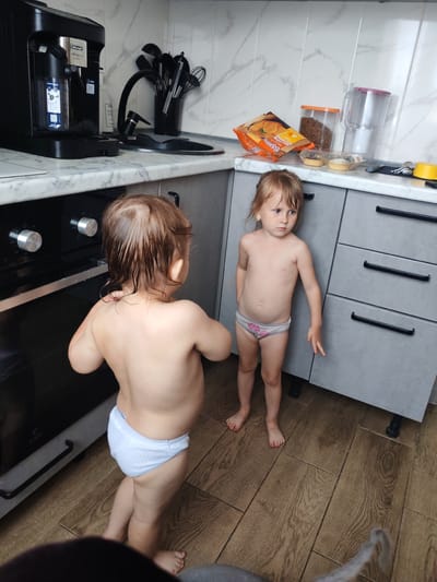 Two young children, a boy and a girl, stand in a kitchen. The boy, seen from behind, is wearing a diaper and has wet hair. The girl, facing away from the boy, is wearing patterned underwear. They are standing in front of a modern gray kitchen counter with a black oven.
On the counter, to the right of a black coffee machine, a collection of kitchen utensils is in a holder. Further to the right, there is a bag of orange snacks, a clear plastic container filled with cereal or chocolate chips, and a water filter pitcher. Two small dessert cups are also on the counter.
The kitchen has light-colored, marble-patterned backsplash tiles and gray cabinets. The floor is made of wood-look tiles. The overall lighting suggests it is daytime. The children appear calm, with the girl looking slightly off-camera with a neutral expression. The location context is Mezhdurechensk, Russia. Visible text on the coffee machine includes "DeLonghi" and "MAGNIFICA EVO".
