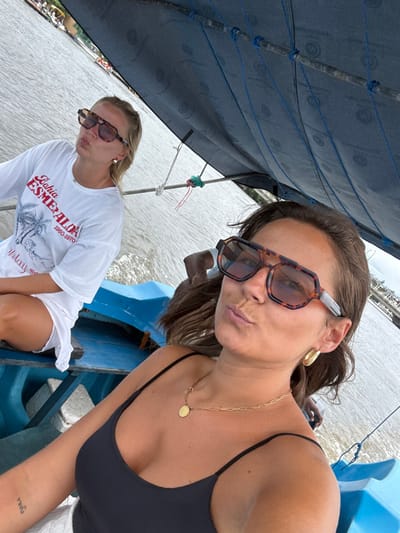 Two women are on a blue boat. The first woman, in the foreground, wears a black swimsuit and stylish sunglasses. A gold necklace adorns her neck. The second woman, seated behind, wears a white T-shirt with a graphic and long sleeves and sunglasses. Both have pouted expressions. A canvas shade provides cover, with blue ropes securing it. The background shows calm water, possibly a river or bay, with distant structures.