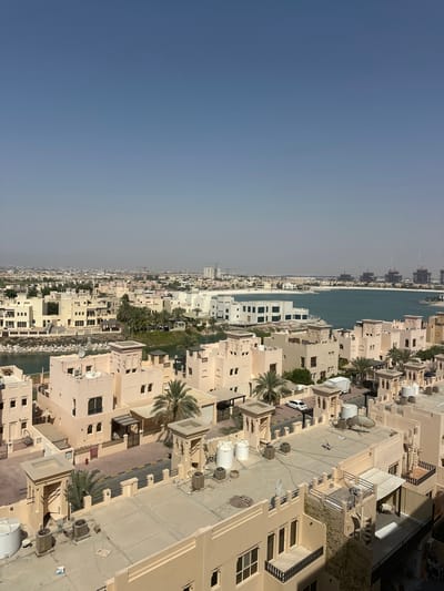 This media file captures an aerial view of a coastal residential area in the United Arab Emirates, likely during daytime. The scene is dominated by a dense arrangement of tan-colored buildings, appearing to be townhouses or villas, with flat roofs. Many of these structures feature multiple windows and balconies. Scattered throughout the development are palm trees, indicative of a warm climate.
The buildings are clustered along a waterway or canal, which separates the residential area from a strip of land that appears to be a beach or promenade, beyond which lies the open sea. In the distance, several tall buildings and construction cranes suggest ongoing development or a more urbanized area further along the coast. The sky is a clear, light blue, transitioning to a hazy or slightly overcast condition towards the horizon, indicating a possibly warm and humid day.
The presence of numerous air conditioning units on rooftops and external walls suggests the need for climate control in this region. Some of the buildings have satellite dishes and water tanks visible on their roofs. A white car is parked on a paved street within the complex. There is no visible text in the image. The overall impression is one of a developed, modern coastal community in a sunny climate.