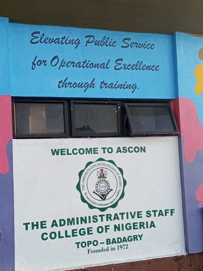 The image shows a painted wall with several text elements, windows, and some painted decoration.
The top section of the wall, painted in blue, reads "Elevating Public Service for Operational Excellence through training" in a darker, cursive font. Below this are three small, dark-framed windows. One of the windows is open slightly.
The bottom section of the wall is painted white. The text "WELCOME TO ASCON" is displayed prominently in green. Below it is the seal of the Administrative Staff College of Nigeria, including the text "THE ADMINISTRATIVE STAFF COLLEGE OF NIGERIA". Underneath, it reads "TOPO - BADAGRY Founded in 1972".
The wall also has a colorful painted decoration in the margins that is a combination of pink, purple and yellow. The image suggests that the Administrative Staff College of Nigeria is located in Topo-Badagry.
