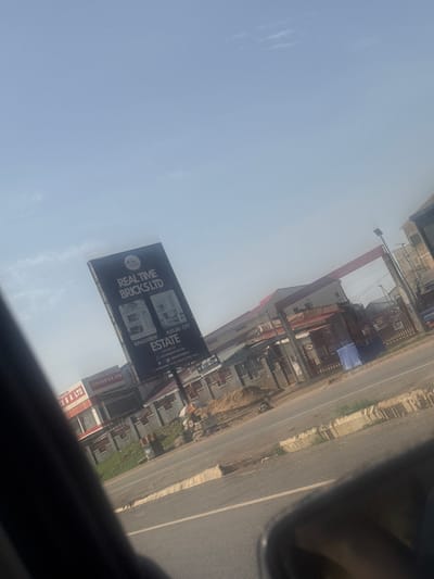 The image depicts a street scene in Kugbo, Nigeria, under a clear sky with scattered clouds. A prominent billboard for "REALTIME BRICKS LTD" advertising "KINGSVIEW ESTATE" and "FUTURE CITY ESTATE" is angled towards the viewer. Adjacent to the billboard, a building with the sign "SIRDIICK G. A. LTD" is visible. The foreground shows a section of a road with a concrete barrier and vegetation along its edge. The background features more buildings, some with red roofing, and what appears to be ongoing construction with piles of sand and materials. There are a few individuals visible, seemingly engaged in work or passing by. The overall impression is one of urban development and commercial activity.