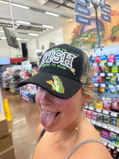An indoor, eye-level photograph depicts a young individual, appearing female, posing within a commercial establishment in Shingle Springs, United States. The individual is positioned centrally, wearing a black and camouflage baseball cap with "BORN TO FISH" and "FORCED TO WORK" embroidered in white and green text, along with a bass fish emblem on the brim. Her face is partially obscured by the cap, and her mouth is open with her tongue fully extended. She wears a grey tank top and a gold-colored chain necklace. The background features store aisles with various products displayed on shelves. Visible overhead are fluorescent lighting fixtures, a hanging black display screen, and a department sign indicating categories such as "HABA," "Pet Food," "Charcoal," "Condiments," and "Pasta." A large wall mural depicting a natural landscape is partially visible on the far wall. A yellow wet floor sign and stacked cardboard boxes are present in the left foreground.