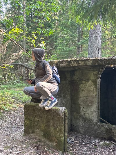 This image shows a person in a crouched position on a moss-covered concrete block within a forest. The subject, wearing a dark hooded jacket, grey pants, and blue and white sneakers, appears to be looking down at a smartphone or small object held in their hand, or into an opening of the concrete structure. A dark backpack is visible on their back.
The surrounding environment is a dense forest, with tall trees featuring both needle-like (coniferous) and broad (deciduous) leaves, indicating a mixed woodland. Bright daylight illuminates the scene, filtering through the tree canopy. The ground is covered with fallen leaves and dirt.
The concrete structure is old and weathered, significantly covered in green moss, suggesting it has been abandoned or unused for a considerable time. It appears to be part of a larger ruin or foundation, possibly a bunker, with a dark, open cavity visible above the person. The overall impression is one of exploration or discovery within a natural and possibly historical setting. No legible text is visible. This scene is set in Komarovo, Russia.