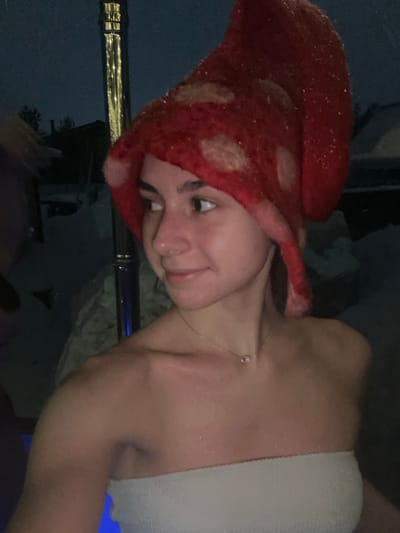 The image displays a young female outdoors at night in Базарово, Russia, likely in a hot tub or warm pool. She is captured from the chest up, facing slightly right with her head turned, looking towards the right with a subtle smile. She wears a red, felted, mushroom-shaped hat with lighter, circular spots and a white bandeau-style top. Her skin appears wet and glistens with water droplets. A delicate silver-colored necklace with a small rectangular pendant is visible around her neck. The background is dark, revealing snow-covered ground and faint outlines of structures. A tall, reflective pole, possibly a heater or light fixture, is visible behind her, emitting small points of yellow light. A blurred, dark mass, potentially another individual or object, is partially visible on the far left. The scene suggests a leisure activity in a cold, outdoor, winter environment.