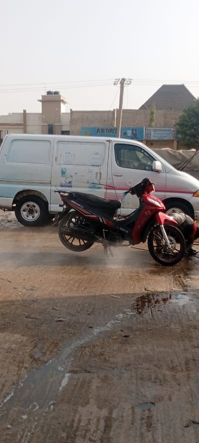 This outdoor scene, set in Kabuga, Nigeria, depicts the washing of a motorcycle. In the foreground, a red and black motorcycle is positioned on a stand, with visible water spray or steam around its engine area, indicating it is actively being cleaned. The lower legs and feet of a person, likely the individual performing the wash, are partially visible near the front wheel. The motorcycle has the text "CARLITO-90" on its side fairing.

Behind the motorcycle, a white cargo van is parked, showing signs of use and faded graphic designs on its side, including faint text like "Active" and "CAFE". The ground beneath both vehicles is wet and reflective in areas, with puddles and streaks of water from the cleaning process.

The background reveals several buildings under a bright, clear sky, suggesting a sunny daytime setting. A prominent sign on one building clearly displays "ABI IYAYE". Next to it, another sign reads "M. Ghaniu Aluminum and Steel Work". A utility pole with power lines is also visible on the right side, framing the busy, everyday environment.