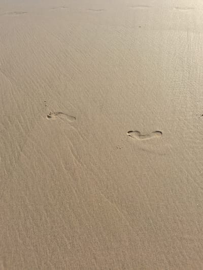 The image shows a sandy beach in Figueira da Foz, Portugal. There are two visible footprints in the sand, indicating a person or persons walked there. The sand is beige in color. The light suggests it may be during the day. The sand shows some subtle ripples on the surface.