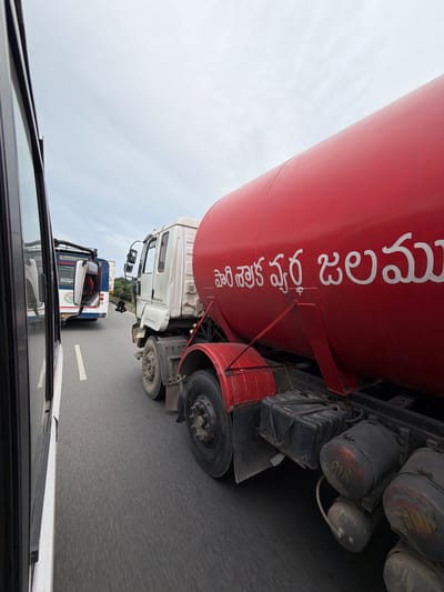 The image captures a scene on a highway in or near Chinna Chilkamarri, India. In the foreground, a white truck with a large red cylindrical tank is prominently visible, traveling on a paved road. The side of the red tank displays white text in Telugu script, which reads "పారిశ్రామిక నీటి శుద్ధి" (Pāriśrāmika nīṭi śud'dhi), meaning "Industrial Water Treatment". To the left of the truck, a white vehicle, likely a bus, is also on the highway, seen from its side. Further in the background, another bus with blue and white markings is visible in traffic. The sky is overcast with clouds. The overall scene depicts highway traffic with commercial vehicles.