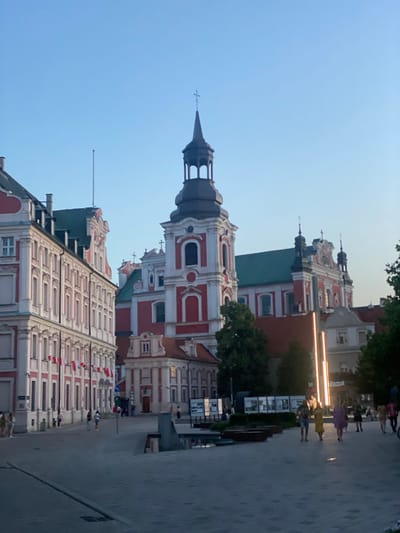 The image captures a view of the Old Market Square (Stary Rynek) in Poznan, Poland, during the late afternoon or early evening. The square is paved with cobblestones and features a fountain area in the foreground. Several people are walking across the square, some appearing to be tourists.

Dominating the scene are the historic buildings surrounding the square. On the left, a long, ornate building with a red and white facade and numerous windows is visible. Flags of Poland hang from this building. In the center, a prominent red and white church with a tall, dark spire and several smaller domes rises against the clear blue sky. To the right of the church, another red and white building with green rooftops and architectural details can be seen.

In the background, modern elements include a row of illuminated vertical lights and a display area with posters or exhibits. The lighting suggests the sun is low in the sky, casting a warm glow on some parts of the buildings while leaving others in shadow. Trees are also present, adding greenery to the urban landscape. The overall atmosphere is one of a bustling European city square.