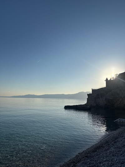 The image shows a coastline in Grad Rijeka, Croatia. The scene includes a rocky cliff face on the right side, a narrow beach with pebbles or small rocks, and calm blue water extending towards the horizon. In the background, there are other landmasses or islands visible. A building or structure is visible on top of the cliff. The sun is shining brightly behind the cliff, creating a lens flare effect. A contrail from a jet is visible across the sky.