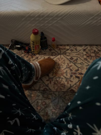 A foot in a brown slipper rests on a patterned rug beside a bed. Beside the slipper is a Coors bottle, a dark bottle, a small clear bottle with a red cap, and a set of keys. The person wearing the slipper is also wearing patterned pajamas. The bed has white linens.