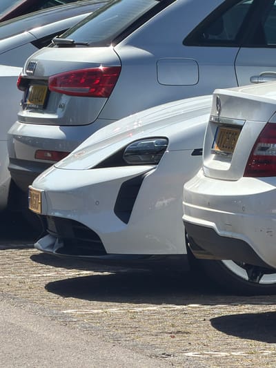 The image depicts a close-up view of two white vehicles parked side-by-side, likely on a street. The focus is primarily on the rear of the vehicles. The vehicle on the right has a visible license plate that reads "VV 2505." The environment appears to be a typical urban setting, with pavement visible to the side of the cars. Based on the prompt, the location is Fentange, Luxembourg. There are no people visible in the frame.