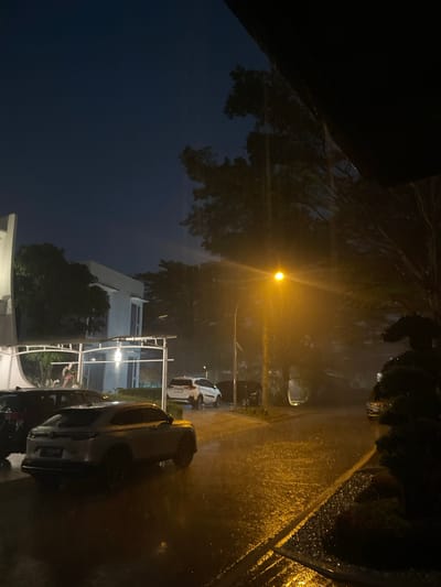 In Babakan Madang, Indonesia, an outdoor scene is depicted during a heavy downpour at night. The foreground features a light-colored SUV, partially obscured by rain, parked on a wet, reflective asphalt road. To the left, a portion of a white building with modern architecture is visible, along with a covered carport structure. Further down the road, a bright yellow streetlamp illuminates the falling rain, creating a hazy glow. Several other vehicles, including a white SUV and a dark-colored car, are parked or driving in the distance. Dense foliage and trees frame the right side of the image, their branches dark against the night sky. The overall atmosphere is dark, wet, and atmospheric, indicative of a significant rainfall event.