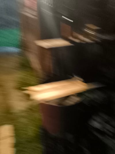 The image is a blurred, horizontal composition that appears to depict an outdoor scene, possibly in the city of Kashino, Russia. Two horizontal, light-colored rectangular objects are visible, suggestive of shelves or planks, one positioned above the other. The upper object is more clearly defined, showing a wood grain texture. The background is a dark, indistinct blur, with hints of green and brown, potentially representing foliage or ground cover. Streaks of light are present, particularly around the upper object, indicating motion blur or a light source. The overall impression is one of movement, making it difficult to identify specific individuals or precise details of the environment.