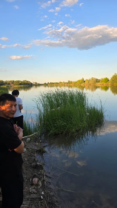 This is a portrait-oriented, low-angle, outdoor shot of two men fishing on the bank of a river at dusk. The sky is a pale blue with scattered, wispy clouds that are catching the warm tones of the setting sun. The river's surface is calm and reflects the sky and the trees lining the opposite bank, creating a mirror image. Tall, lush green grass grows from the bank of the river, with some sparse white flowers visible within it. The foreground is dominated by the muddy, uneven bank, littered with debris including plastic bags and branches.
The man in the foreground is wearing a black t-shirt and is lying on his side, looking towards the camera. His face is partially illuminated by the warm sunlight, highlighting a relaxed and perhaps contemplative expression. The second man, further back and wearing a white t-shirt, is also facing away from the camera, appearing to be focused on fishing. A fishing rod is visible near him, and another is resting on the bank in the foreground. The overall atmosphere is peaceful and serene, suggesting a quiet evening spent in nature. The time of day is clearly dusk, indicated by the low angle of the sun and the warm, golden light. The location is suggested to be Uralsk, Kazakhstan, which is consistent with the natural landscape depicted. No text is visible in the image.