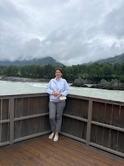 A woman stands on a wooden deck in front of a railing, with a river and mountainous landscape in the background. She is wearing a light gray hooded sweatshirt, patterned black and white pants, and silver slip-on shoes. She has a white purse slung over her shoulder. The river is wide and appears to be flowing rapidly, with rocky outcrops visible in the water. The mountains are covered in dense green forest and are partially obscured by clouds. The sky is overcast. The location is identified as Elekmonar, Russia.