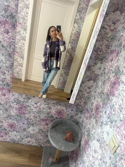 A young woman with dark hair pulled back in a ponytail is taking a mirror selfie. She is wearing a purple and white plaid jacket over a dark top and light-wash jeans. She is standing in what appears to be a room with floral wallpaper and a wooden floor. To her right, a full-length mirror reflects the scene. Partially visible in the reflection is a white door. In the foreground, a grey cat tree with a plush toy is visible.