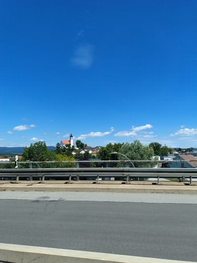 A wide shot captures a bright, clear blue sky with a few scattered white clouds. In the mid-ground, a small town is nestled amidst lush green trees. A prominent white church with a dark spire and a red-tiled roof stands out on a small hill. The town features various buildings with red and brown roofs. A metal guardrail runs horizontally across the foreground, separating the viewer from the town and sky. Below the guardrail, a section of a road is visible, with trees and foliage peeking out from behind it. In the far background, rolling hills are visible under the bright sky. The image appears to be taken from a bridge overlooking the town. The scene is tranquil and bathed in natural sunlight.