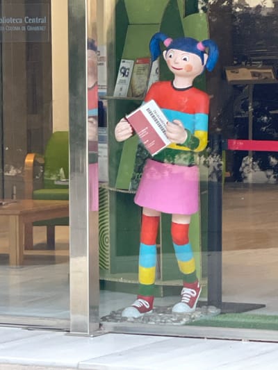 The image shows a colorful statue of a young girl with blue pigtails, wearing a striped shirt, pink skirt, and striped socks, holding a book. The statue is positioned in front of a display in what appears to be a library or community center, indicated by the text "Biblioteca Central" and "STA. COLOMA DE GRAMENET" visible on a sign in the background, which places the location in Santa Coloma de Gramenet, Spain. The environment is brightly lit, and the statue is surrounded by shelving units and a green chair is visible in the background through a glass pane.