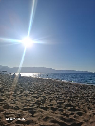 A sunlit beach scene, possibly near Gazi Municipal Unit, Greece. The foreground is dark, sandy beach, with prominent footprints. The sea is visible, with the sun reflecting on the water surface. In the background, distant mountains are silhouetted against the horizon. The sky is clear blue, with the sun shining brightly. A group of people are distantly visible near the horizon line on the sand. The text "Galaxy A55 5G" is written in the lower right corner.