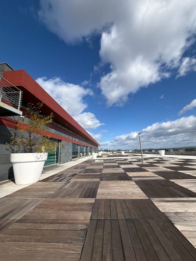 This image depicts a rooftop terrace in Viseu, Portugal, under a clear blue sky dotted with scattered white cumulus clouds. The foreground features a checkerboard pattern of rectangular wooden planks, alternating between lighter and darker tones. To the left, a modern building extends horizontally, presenting a facade composed of a dark grey lower section with large rectangular windows, topped by a prominent red horizontal band. A metal-railed structure, possibly part of a balcony or external staircase, is visible on the far left side of the building.
Along the building's edge and extending across the terrace, several large, white, conical planters are positioned. The planter in the immediate foreground contains a small deciduous tree displaying yellow-orange autumn foliage. Multiple tall, slender, silver-colored lampposts are evenly spaced across the wooden flooring towards the right and in the background. The far edge of the terrace reveals a low railing or parapet overlooking a distant, somewhat hazy cityscape or landscape. No individuals are present in the scene. The daylight is bright, casting visible shadows.