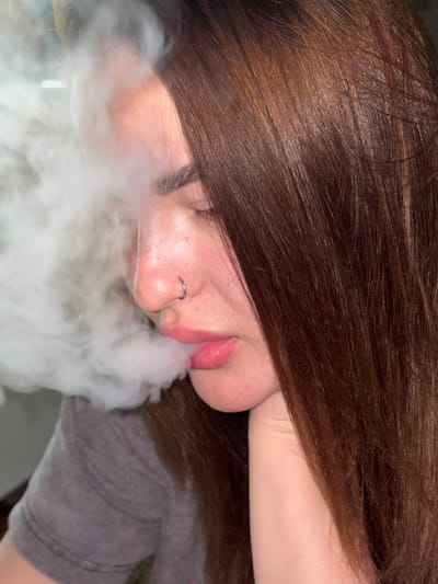 A close-up, angled shot depicts a person exhaling a large cloud of vapor. The individual has brown hair that partially obscures their face. A silver ring is visible in their left nostril. The person is wearing a gray t-shirt. The location is not discernible from the provided image.
