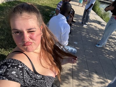 An outdoor, high-angle, eye-level selfie shows a young woman sitting on a grassy area with a paved walkway behind her and a body of water to her right. The woman is looking towards the camera with her right eye closed and her lips pursed. She has long, brown hair that is partially obscuring her face and is wearing a black and white patterned off-the-shoulder top. She has a nose ring and earrings. Several other individuals are visible in the background, including a person in a white shirt and grey pants, and another person in a blue top and shorts. The environment appears to be a park or recreational area, possibly along a waterfront. There are shadows from trees on the paved walkway. The image appears to have been taken during daylight hours. The context of Weil am Rhein, Germany, is not visually evident within the image itself, though the setting suggests a public outdoor space.