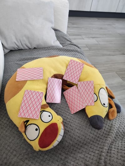 An indoor scene depicts a yellow plush toy, shaped like a cartoon dog with a wide open mouth and bulging eyes, lying on a gray textured blanket. Several playing cards, with their red and white checkered backs facing up, are scattered across the toy and the blanket. One card is partially obscured beneath the toy's head. Behind the toy, a light-colored pillow rests on the blanket. The background shows a wooden floor with a matte finish and a section of kitchen cabinets. There is no information in the image to indicate a location in Grosseto, Italy.