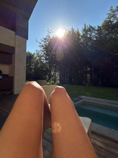 The image shows a person's legs, tanned from the sun, resting in a lounge chair on a wooden deck. The deck is adjacent to a small, tiled swimming pool. Behind the pool, a lawn is visible, bordered by a dense forest. The sun is high in the sky, creating a bright glare and lens flare. To the left of the deck is a modern building with stone and wood accents. The setting appears to be a private residence in a natural, wooded area. While the city of Bakovići, Montenegro is mentioned in the prompt, there are no specific visual cues within the image to confirm this location.