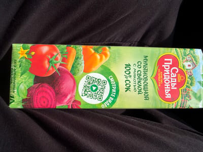 The image shows a close-up, top-down view of a juice box lying on a dark, textured fabric, likely clothing. The juice box is rectangular and oriented horizontally, with its packaging prominently displayed. The dominant colors on the packaging are green, red, and yellow, depicting a variety of vegetables.
On the left side of the box, there is a red heart-shaped logo with yellow and white trim, featuring a pastoral scene with a house and trees. Inside the heart, text in red and white reads "Сады Придонья," and below that, in smaller white text, "КАЧЕСТВО ИЗ ПЕРВЫХ РУК."
To the right of the logo, larger text in white and green states "МУЛЬТИОВОЩНОЙ СО СВЁКЛОЙ С МЯКОТЬЮ 100% СОК." In the center, a circular emblem contains a QR code and the Russian phrase "СМОТРИТЕ ВИДЕО" (Watch video).
The right side of the juice box features illustrations of vegetables: a large beet, a bell pepper, a cucumber, a carrot, and a tomato with a small yellow flower. Along the far right edge, vertical text in green reads "Без добавления сахара" (Without added sugar).
The lighting suggests it is daytime, with a bright light source creating some shadows and highlights on the fabric and the juice box. The overall impression is a product shot, emphasizing the natural ingredients and juice variety. The location context is Нуркеево, Russia.