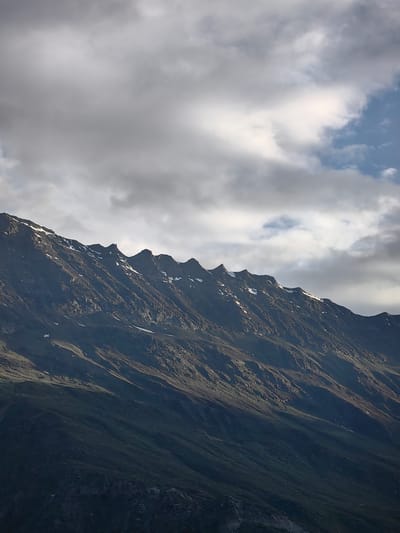 This media file is a vertical, eye-level, medium shot of a rugged mountain range under a cloudy sky. The mountains are dark and rocky, with streaks of snow visible on their upper slopes. The peaks are sharp and jagged, creating a serrated outline against the sky. The lower slopes transition to a gradient of dark green, suggesting vegetation or shadowed terrain. The sky is filled with a mix of heavy, gray clouds and patches of blue, indicating a dynamic weather system. The lighting suggests it is either early morning or late afternoon, with the sun intermittently breaking through the clouds to cast light on the mountain. There is no discernible activity or event taking place, and no people, objects, or text are visible. The scene evokes a sense of raw, natural beauty and the dramatic grandeur of a mountainous landscape. The location is identified as Stingri, India.