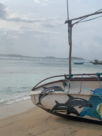 The image shows the bow of a fishing boat with dolphin paintings on its side, resting on a sandy beach in Weligama, Sri Lanka. The ocean is visible in the background with gentle waves and other boats further out. The sky is overcast with clouds. The boat has a wooden mast and is secured with ropes. The beach is wet near the water's edge and dry further up. The overall scene suggests a serene, possibly early morning or late afternoon setting.