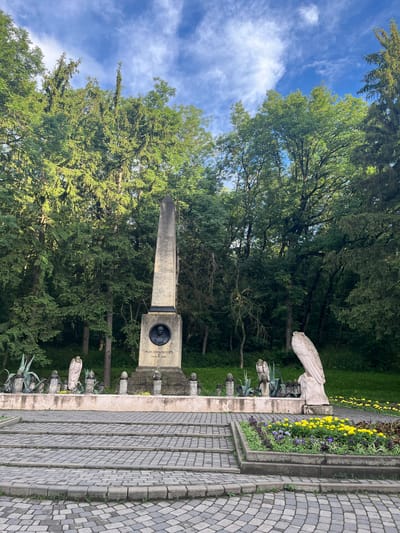 The image shows a monument in Pyatigorsk, Russia, dedicated to Mikhail Lermontov. The monument is an obelisk with a circular plaque featuring a portrait of Lermontov. The obelisk is surrounded by sculptures of mythological creatures, some of which appear to be eagles. In the foreground, there are tiered steps made of cobblestone, leading up to a paved area. A flower bed with yellow and purple flowers is visible on the right side of the steps. The monument is situated in a park-like setting with tall trees forming a dense green backdrop. The sky is blue with scattered white clouds. The scene is static, with no people or animals actively present.