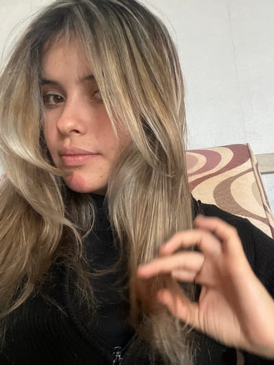 A young woman with long, blonde, highlighted hair is captured in a close-up, frontal shot. She is looking slightly to her left, her brown eyes visible beneath bangs that partially obscure her face. Her lips are closed, and her chin displays a red, irritated patch. She is wearing a black, high-necked garment. Her right hand is raised in front of her chest, with her fingers curved as if holding something small or making a gesture. Behind her, to her right, is the patterned upholstery of a chair, featuring swirling shapes in shades of cream, brown, and reddish-purple. The background wall is textured and light-colored, suggesting an indoor domestic setting. The overall lighting is soft, illuminating her face and hair.