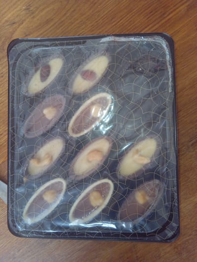 A rectangular, clear plastic package containing twelve oval-shaped chocolates is presented on a wooden surface. The chocolates are arranged in three rows of four. Each chocolate features a lighter-colored topping, possibly marzipan or white chocolate, with a darker chocolate base. Some chocolates have what appear to be nuts or dried fruit embedded in the lighter topping. The packaging is dark with a subtle gold geometric pattern. The wooden surface is a medium brown with visible grain. There is no indication of the location within Terebovlia, Ukraine.