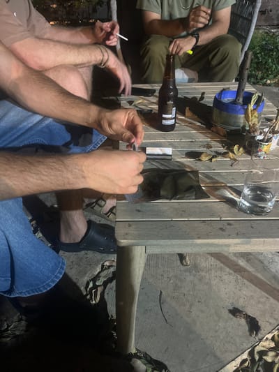The image depicts an outdoor, nighttime scene in Ganei Tikva, Israel, showing three individuals engaged in a casual gathering around a light-colored wooden slatted table.
In the foreground, a person wearing blue denim shorts and a black Croc-style sandal on their left foot is actively rolling a cigarette or joint. Their hands are centered, holding rolling papers and material. To their left, another individual, wearing light-colored attire, is visible with their left arm resting on the table, holding a lit cigarette between their thumb and forefinger. A gold-colored bracelet is present on this person's left wrist. In the background, a third person, dressed in olive green pants and a matching top, is partially visible, with a watch on their left wrist and their hand raised.
On the table, several items are arranged: a dark brown glass bottle, identified as a "Goldstar" beer, stands upright. Adjacent to it, a small, rectangular, white and grey patterned object lies flat. A dark green or black pouch, partially open, contains a quantity of leafy, granular material consistent with cannabis. A clear glass containing water is positioned on the right side of the table. A blue and silver bucket-like container with a vertical metal shaft and dried plant material, possibly indicative of a water pipe, is visible further right. Several small, multi-colored decorative pots containing withered plant matter are also present. Dry leaves are scattered across the table surface. The ground beneath the table is concrete, partially covered with fallen dry leaves. Illumination suggests artificial lighting, casting shadows and highlighting the subjects and objects.