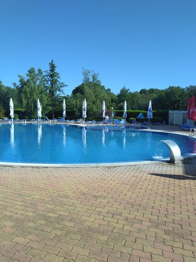 A sunny, clear blue sky hangs over a resort swimming pool in Yagoda, Bulgaria. The pool is filled with bright blue water and bordered by a paved walkway made of interlocking red and yellow bricks. Several white and blue umbrellas stand open around the pool, providing shade for empty lounge chairs. A modern, silver waterfall feature is installed at the edge of the pool, creating a gentle cascade of water. In the background, lush green trees and dense foliage form a natural backdrop. A few structures, likely changing rooms or small buildings, are visible further back. The scene suggests a peaceful and inviting atmosphere for relaxation and recreation.