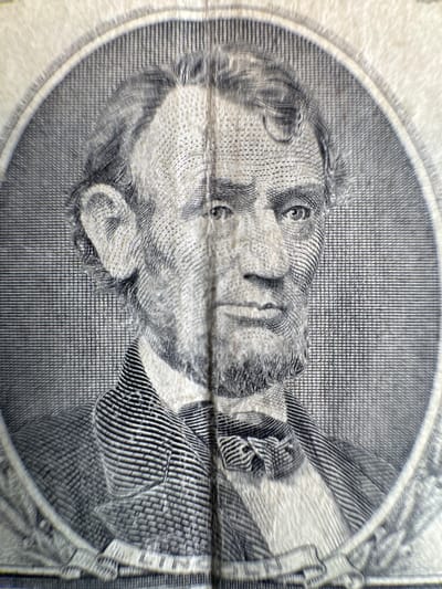 The image is a close-up, macro photograph of Abraham Lincoln's portrait on a U.S. five-dollar bill. The engraving details are highly visible, showcasing the texture of the paper and the intricate lines that form Lincoln's face, hair, beard, and clothing, including his suit and bow tie. The circular border of the portrait is also evident. The lighting is even, highlighting the details of the engraving. The image does not depict any location in the city of Mejaniga, Italy, as it is a photograph of currency.
