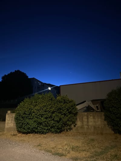A night photograph depicts a view of buildings and vegetation in the city of Anduze, France. The sky is a deep, gradient blue, transitioning from a darker shade at the top to a lighter hue near the horizon. In the upper right portion of the sky, a small, faint point of light, possibly a distant star or planet, is visible.

In the mid-ground, a building with multiple windows, some illuminated from within, stands silhouetted against the sky. To its left, a large, dark tree with dense foliage obscures part of the building. A bright, rectangular light source emits a beam of light from the building, angled downwards and to the right.

In the foreground, a large, rounded, dense green bush dominates the left side of the frame. Behind it, a weathered concrete wall runs horizontally across the image. Further to the right, another, smaller green bush is visible. Peeking over the concrete wall and behind the right-hand bush is a white construction vehicle, possibly a backhoe, with its arm extended. The ground in front of the wall is covered with dry, sparse grass and a gravel path. The overall impression is one of a quiet, possibly residential area at dusk or after dark.