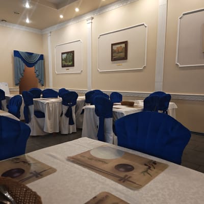 The image captures an empty dining hall or restaurant, meticulously set up for guests. The room features light-colored (beige/cream) walls adorned with ornate white decorative molding, including crown molding and fluted pilasters, creating a classical aesthetic. Several framed landscape paintings depicting forest scenes, one with green trees and another with autumn foliage, hang within these wall panels. Small, unreadable plaques are visible beneath each painting.

The ceiling is also light-colored and fitted with several recessed spotlights providing illumination. In the background, a doorway or archway is framed by elegant blue and grey draped curtains. A white appliance, possibly a water cooler, stands to the left of this archway.

The room is furnished with numerous tables draped in white tablecloths. Each chair is covered in a vibrant royal blue fabric, tied neatly with a bow at the back. On the tables closest to the foreground, brown placemats with Asian-inspired designs, featuring what appears to be a bowl, chopsticks, and green foliage elements, are visible. Some tables also have small woven baskets and white condiment containers. The floor is tiled or concrete, visible beneath the table coverings.

The setting appears formal and ready for an event or meal service, but no people are present. The lighting suggests an indoor evening setting or an area reliant on artificial light. The overall atmosphere is quiet and orderly.